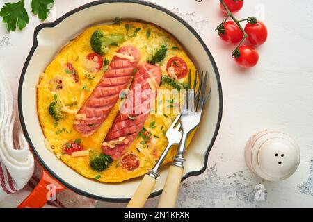 Omelette ou frittata avec deux saucisses, fromage fondu, tomates cerises, brocoli et persil sur poêle rouge ou poêle en fer sur fond blanc en béton Banque D'Images