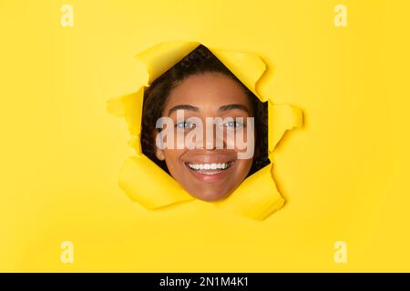 Femme afro-américaine excitée posant sur fond de papier jaune déchiré, regardant la caméra et souriant à travers le trou Banque D'Images