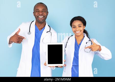 Nouvelle application médicale. Médecins noirs, hommes et femmes, pointant vers une tablette numérique avec écran vide, maquette Banque D'Images