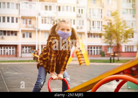 Petite fille avec masque médical sur le terrain de jeu pendant la quarantaine Covid-19 Banque D'Images