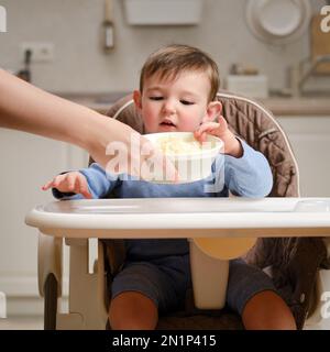 Un enfant drôle mange une pomme râpée assise sur une chaise de cuisine. Bon petit garçon mange de la nourriture. Enfant âgé d'un an et de quatre mois Banque D'Images