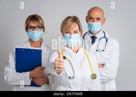 Prise de vue d'une équipe médicale portant un masque facial et se tenant ensemble sur un fond isolé tandis que la femme médecin donne des pouces vers le haut. Banque D'Images