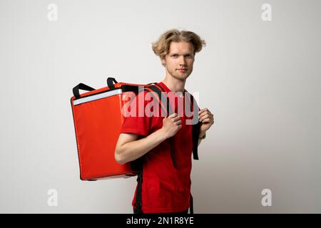 Jeune livreur en uniforme rouge avec sac thermo isolé sur fond blanc. Service de livraison. Banque D'Images