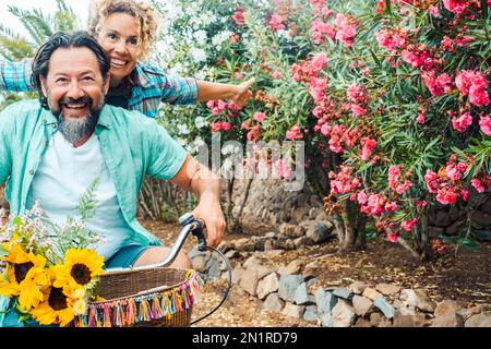 Jeune couple d'âge mûr vous pourrez profiter de loisirs en plein air en vous amusant et en riant. Homme et femme à vélo à l'extérieur avec des fleurs en ba Banque D'Images