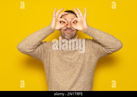 Homme beau barbu portant le turtleneck sur fond jaune isolé faisant un geste ok comme des jumelles, les yeux regardant à travers les doigts. Fou Banque D'Images