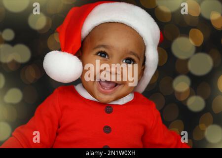 Joli petit enfant afro-américain portant un chapeau de père Noël contre des lumières floues sur fond sombre. Fête de Noël Banque D'Images