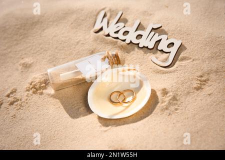 Coquillage avec anneaux dorés, invitation en bouteille de verre et mot Wedding sur la plage de sable Banque D'Images