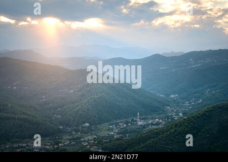 Vue d'été sur la ville de montagne sur la Riviera italienne au coucher du soleil. La lumière du soleil à travers les nuages illumine les montagnes. Ligurie, Italie Banque D'Images