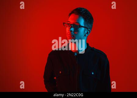 Studio Portrait d'un beau jeune homme de brunette caucasien à la mode portant des lunettes de vue dans une combinaison élégante sur fond lumineux rouge néon Banque D'Images