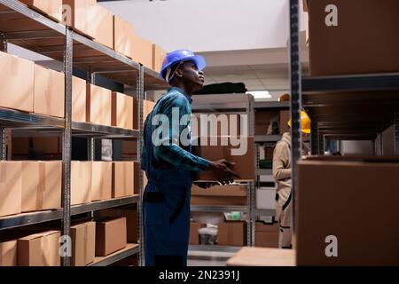 Chargeur de magasin afro-américain contenant une boîte en carton emballée et regardant l'étagère pleine de conteneurs prêts à être expédiés. Employé de l'entrepôt gérant le transport des colis Banque D'Images
