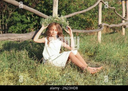 Jolie petite fille portant une couronne faite de belles fleurs près d'une clôture en bois à l'extérieur le jour ensoleillé Banque D'Images