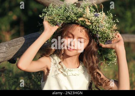 Jolie petite fille portant une couronne faite de belles fleurs près d'une clôture en bois à l'extérieur le jour ensoleillé Banque D'Images