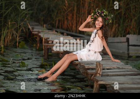 Jolie petite fille portant une couronne faite de belles fleurs sur la jetée près de l'étang au coucher du soleil Banque D'Images