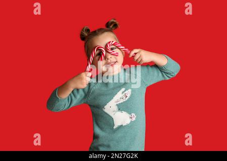 Jolie petite fille en pull tenant des cannes de bonbon sur fond rouge. Célébration de Noël Banque D'Images