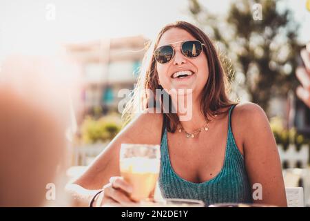 Jeune fille souriante en lunettes de soleil à la table extérieure avec des amis. Boire une boisson fraîche le jour d'été ensoleillé, avec le soleil créant une lumière chaude eff Banque D'Images