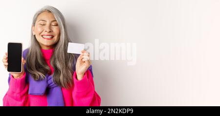 Achats en ligne. Heureuse et ravie asiatique femme senior souriant avec joie, montrant l'écran vide de smartphone et carte en plastique, l'achat dans Internet, blanc Banque D'Images