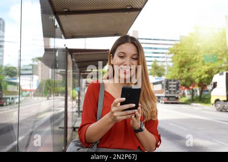 Positive sourire fille achetant le billet en ligne avec smartphone à l'arrêt de bus Banque D'Images