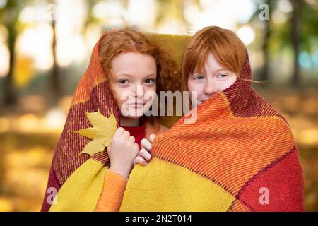 Deux amies aux cheveux rouges se sont enveloppées dans une couverture en laine dans un parc d'automne. Les enfants en automne. Banque D'Images