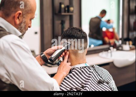 Vue arrière du crop barber tailler les cheveux sombres de la cliente dans le barbershop moderne pendant la journée de travail Banque D'Images