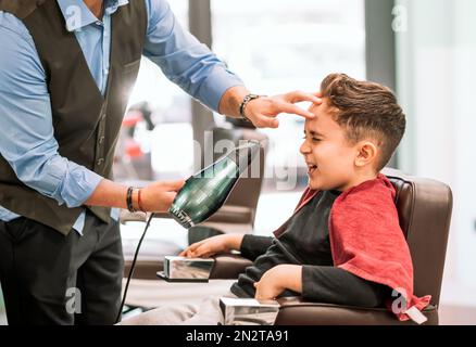 Vue latérale de crop anonyme barber en uniforme séchage des cheveux des enfants assis sur un fauteuil dans un salon de coiffure moderne Banque D'Images