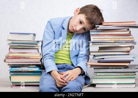 Un garçon fatigué d'âge scolaire est assis entre des piles de livres. Triste enfant avec des livres. Banque D'Images