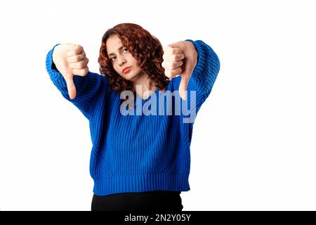 Portrait d'une femme mignonne portant un pull-over se tenant isolé sur fond blanc fait un geste de pouce vers le bas avec les deux mains. Pas satisfait et Banque D'Images