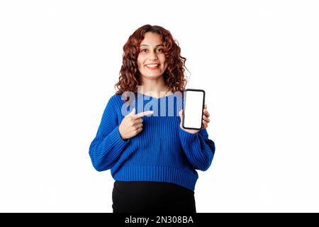 Portrait d'une jeune femme portant un pull-over bleu isolé sur fond blanc tient le téléphone avec la main et pointe avec l'index vers l'emp Banque D'Images