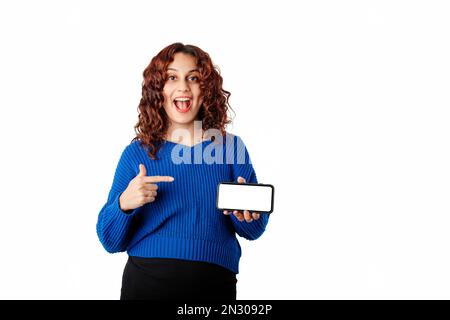 Portrait d'une jeune femme debout isolée sur fond blanc tenant le téléphone horizontalement à la main, pointe vers l'écran blanc maquette avec index Banque D'Images