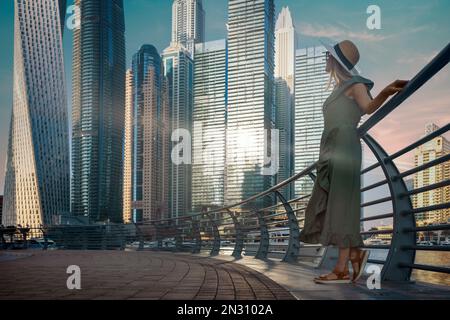 Femme touristique vêtue d'un chapeau et d'une robe d'été tendance et profitant de l'horizon de la marina de Dubaï au coucher du soleil. copier l'espace Banque D'Images