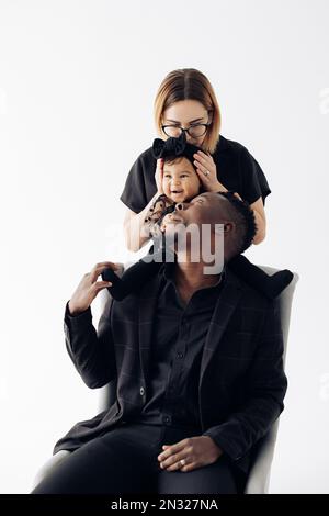 Bonne famille interraciale avec bébé fille a du plaisir et rires près d'un fauteuil sur fond blanc. Gros plan. Concept de famille et d'unité interraciales Banque D'Images