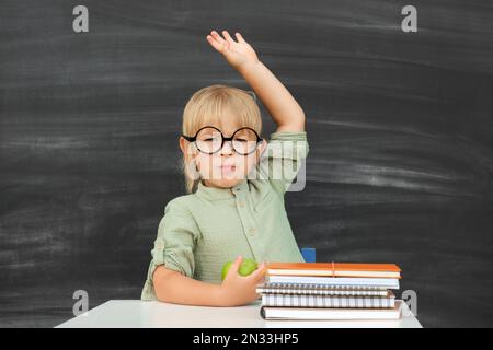 Retour à l'école. Joyeux élève souriant dans des verres ronds assis au bureau. L'enfant lève la main dans la salle de classe avec le tableau noir sur fond. Enfant Banque D'Images