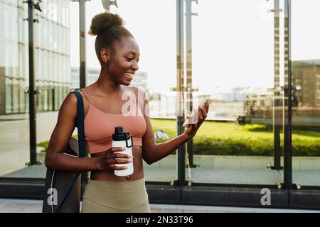 Jeune belle femme souriante afro avec bouteille et tapis tenant et utilisant son téléphone, tout en se tenant à l'extérieur Banque D'Images