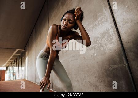 Jeune femme africaine fatiguée sportive dans un casque essuyant son front avec les yeux fermés , tout en se penchant sur les genoux debout à l'intérieur Banque D'Images