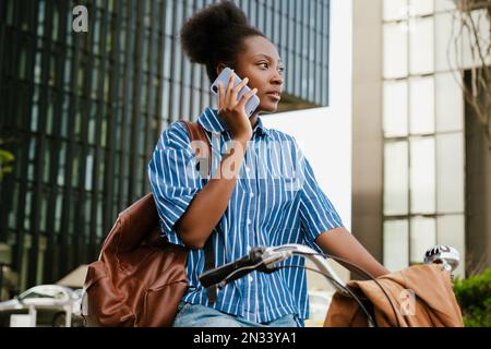 Portrait d'une jeune femme africaine belle parlant téléphone et regardant de côté en se tenant debout avec vélo à l'extérieur Banque D'Images