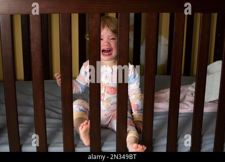 Petite fille caucasienne pleurant dans le berceau en pyjama. Triste enfant malade. Enfant qui pousse les dents Banque D'Images