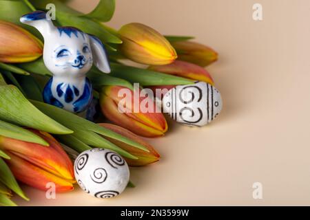 Joyeuses Pâques ! Œufs décorés, tulipes rouges et lapin en porcelaine sur fond beige. Banque D'Images