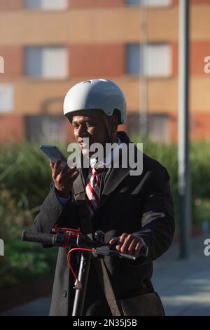 Jeune homme d'affaires latin conduisant une moto électrique dans la rue de la ville et parlant au téléphone. Banque D'Images