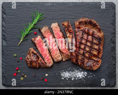 Délicieuses tranches de steak de ribeye grillées et quelques assaisonnements sur une assiette de service en ardoise noire. Pose à plat. Banque D'Images