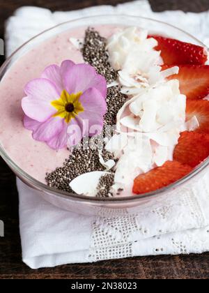 Bol à smoothie au yogourt aux bananes et aux fraises, garni de fraises en tranches, de graines de chia, de flocons de noix de coco et de fleurs comestibles de primula Banque D'Images