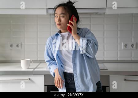 Femme asiatique pensive fatiguée debout dans la maison à la cuisine avec une tasse de café faisant appel dans le téléphone portable Banque D'Images