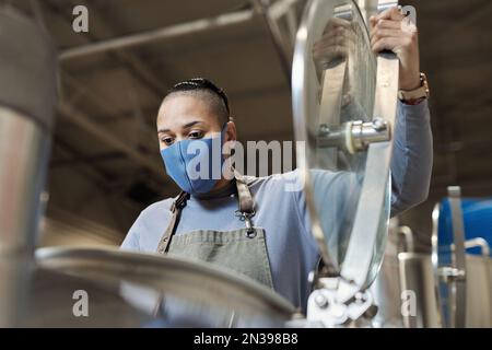 Portrait à angle bas de la brasseur femelle portant un masque ouvrant le réservoir à l'usine de fabrication de bière Banque D'Images