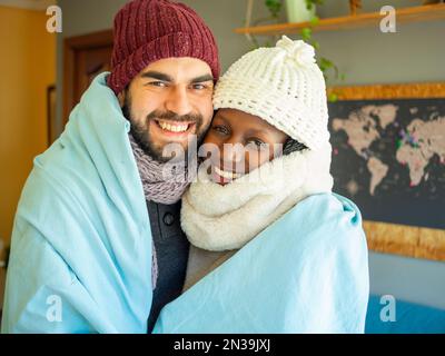 Un jeune couple partage une couverture pour combattre la vague de froid. Porter des vêtements d'hiver dans un salon confortable. Regarder l'appareil photo. Banque D'Images