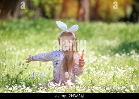 Petite fille portant des oreilles de lapin de pâques gnaws carotte. Joyeuses pâques. Vacances de printemps. Fanny enfant blond dans le jardin de printemps, fleur blanche en fleurs Banque D'Images