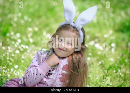 Portrait de fille souriante avec de longs cheveux blonds joue sur le champ de camomille, à l'extérieur. Enfant s'amusant sur la chasse aux œufs de Pâques. Adorable petite fille porte bu Banque D'Images