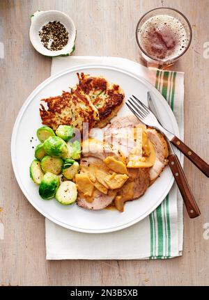 Rôti de porc avec sauce aux pommes, choux de bruxelles et galettes de pommes de terre sur une assiette blanche avec fourchette et couteau Banque D'Images