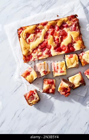Carrés de rhubarbe avec des couches de fudge et de fromage à la crème coupées en morceaux sur papier parchemin blanc sur fond de marbre Banque D'Images