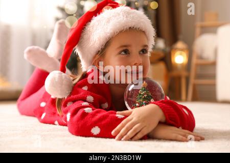 Petite fille dans chapeau de père Noël jouant avec boule de neige sur le sol Banque D'Images