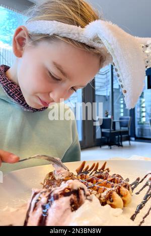 Une adolescente dans un cerceau de lapin mange une délicieuse gaufre avec du sirop de chocolat et de la crème glacée dans un café. Concept de temps familial Banque D'Images