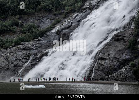 Les touristes observent le flux puissant des chutes Nugget à la base de Bullard Mountain en aval du glacier Nugget. La chute d'eau descend de 377 pieds sur... Banque D'Images