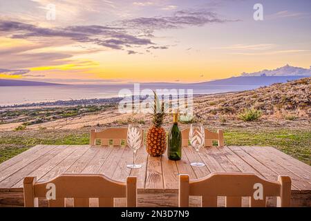 Ananas, bouteille de vin et deux verres à vin assis sur une table en bois avec des chaises sur une colline surplombant Lauuniupoko et l'océan Pacifique... Banque D'Images